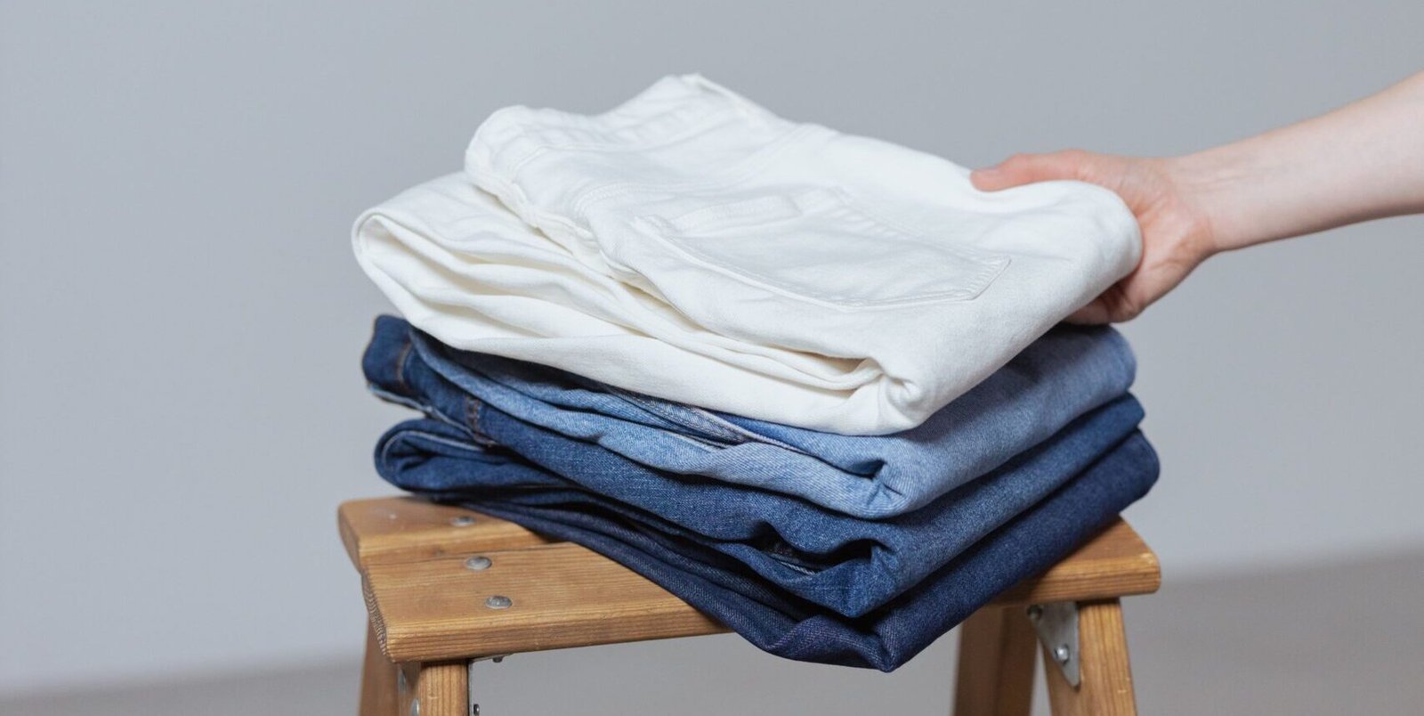 A hand holding a stack of neatly folded white and blue jeans against a minimalist gray background.