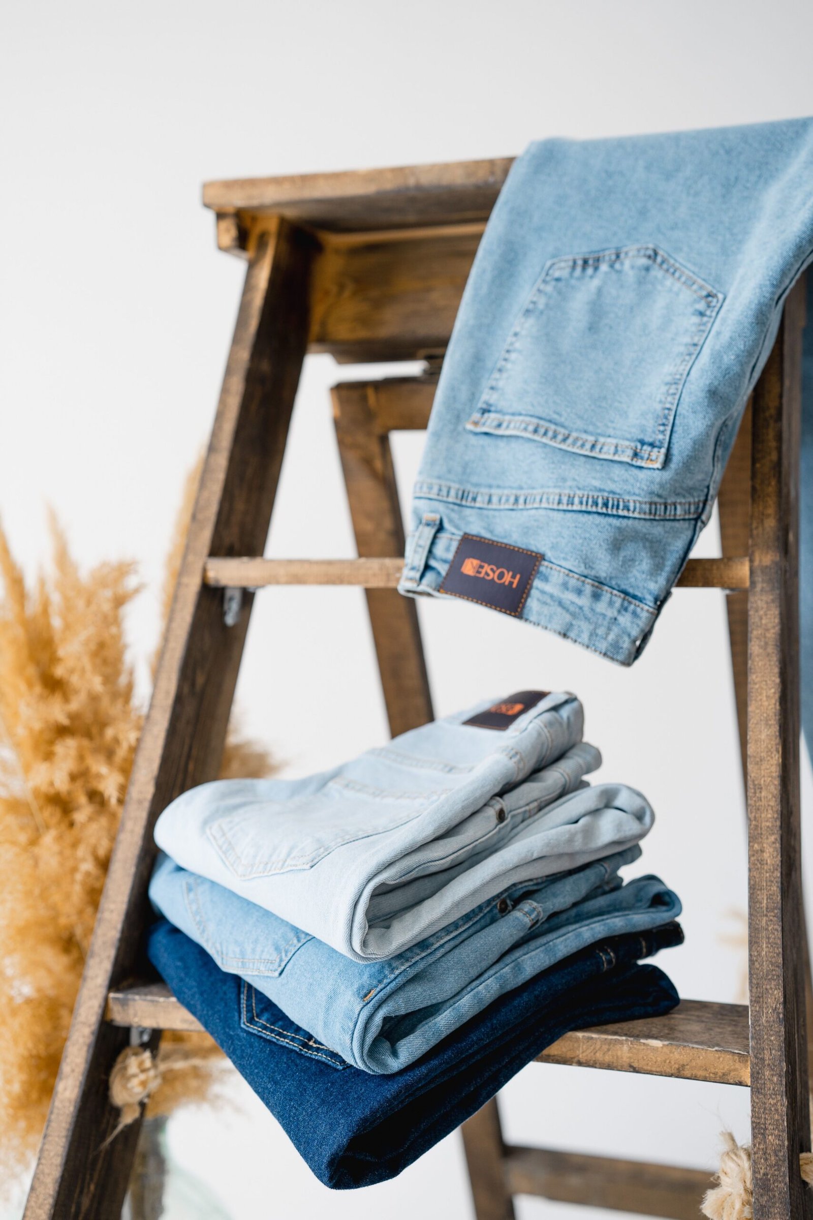 Light blue jeans draped over a wooden stool above a stack of folded denim in various blue washes.